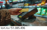 A hand holds a yellow card near a payment terminal on a marble countertop. The background shows shelves stocked with various products. Стоковое AI-видео, аниматор Nart / Фотобанк Лори