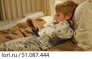 Cute kid boy in pajamas lying in bed watching smartphone, smiling. Children's screen time. Child plays games using phone at the evening or night. Social media addiction. Childhood, modern technology. Стоковое видео, видеограф Марина Демидюк / Фотобанк Лори