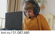 Little smiling 6 years old boy using a laptop with headphones. Child having video call with friends, granny at home in bed. Kid looking at display and waving hand. Childhood leisure activity concept. Стоковое видео, видеограф Марина Демидюк / Фотобанк Лори