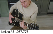 Senior man is playing guitar. Elderly man sitting on the sofa and playing guitar. Portrait of a gray-haired mature man in a sweater and glasses learning to play. Enjoying retirement life at home. Стоковое видео, видеограф Марина Демидюк / Фотобанк Лори