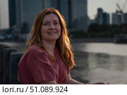 Woman with long hair smiling in warm sunset light, sitting by the river with modern skyscrapers blurred in the background. Стоковое фото, фотограф Софья Соломонова / Фотобанк Лори