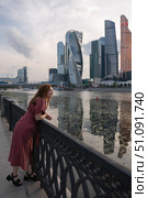 Woman in a red dress leaning on a riverside railing with Moscow City skyscrapers and reflections in the water at sunset. Редакционное фото, фотограф Софья Соломонова / Фотобанк Лори