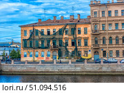 A building in the historic center of Saint Petersburg, currently undergoing renovation or showing signs of neglect. The facade is draped with protective netting, some windows are broken, and the plaster is deteriorating. In the foreground is an embankment and a canal or river. The image captures the contrast between cultural heritage and the passage of time — a typical scene in the older districts of Saint Petersburg, rich with charm and historic character. (2017 год). Редакционное фото, фотограф Анна Кривицкая / Фотобанк Лори