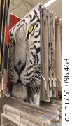 Carpet and flooring store. The center of the frame features a carpet with a bright and striking design of a white tiger's head with yellow eyes. The black and white image of the predator creates a contrasting and expressive impression, making the product a decorative accent. The carpets are hung vertically for display, with other models with traditional and modern patterns in calm tones visible nearby. Rolled carpets and rugs are located on a shelf below. The scene conveys the atmosphere of the home interior department in a home goods hypermarket. (2019 год). Редакционное фото, фотограф Анна Кривицкая / Фотобанк Лори