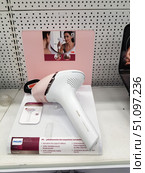 Philips Lumea Prestige IPL photoepilator, displayed in the store on a demonstration stand. The device has a stylish modern design: a white body with pink and gold inserts. In the background is an advertising poster with an image of a woman using the device to remove facial hair. The stand lists the main characteristics of the model: long-term smoothness of the skin up to 8 weeks, reduced irritation and ingrown hairs, 40% higher speed compared to previous versions, the ability to use both with and without a wire. The atmosphere of the photo conveys a sense of modern self-care, beauty and innovative technologies. (2019 год). Редакционное фото, фотограф Анна Кривицкая / Фотобанк Лори