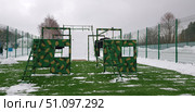 Outdoor sports and training ground in winter. In the center of the frame is a military-style obstacle course, painted in green camouflage with yellow-brown spots. The structure is made of a metal frame and includes ladders, windows for climbing through, and suspended car tires used as training elements. In the background is a high white wall for storming and overcoming. The area is enclosed by a high green fence covered with mesh. The ground is covered with artificial turf, covered in snow in places. The atmosphere is wintry, the sky is cloudy, the trees in the distance are leafless. Стоковое фото, фотограф Анна Кривицкая / Фотобанк Лори