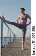 Woman stretching leg on metal railing while standing on concrete pier by the sea under morning sunlight, front view fitness routine improving flexibility, balance and active healthy coastal lifestyle. Стоковое видео, видеограф Сергей Тимофеев / Фотобанк Лори