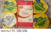 A set of cheeses in factory packaging. In the center is a large rectangular piece of Milbona brand Edam cheese weighing 700 grams in red and white packaging with an image of sliced cheese on the front. Around it are four round packages of Finnish Valio Oltermanni cheese in yellow packaging with a green logo and the Finnish flag. Three of them are a light version of Kevyempi 17%, one is the classic Alkuperäinen. The cheeses are lying on a soft surface, which creates a feeling of home comfort and buying groceries for the family. The photo conveys the brightness of the packaging and the variety of the assortment well, emphasizing the quality of European products. (2019 год). Редакционное фото, фотограф Анна Кривицкая / Фотобанк Лори