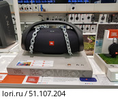 Display cases in an electronics store with a portable JBL Boombox speaker. The large black speaker system with the brand logo is placed on a display stand and secured with a metal chain for protection. Nearby are promotional materials describing the characteristics: Bluetooth, up to 24 hours of playback, IPX7 waterproofness, powerful stereo sound. In the background, you can see other products - headphones, speakers and accessories from various brands. The photo emphasizes the modern style and popularity of audio equipment for home and outdoor activities. (2019 год). Редакционное фото, фотограф Анна Кривицкая / Фотобанк Лори