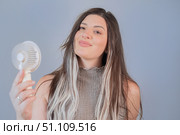 Young woman smiling while holding a portable mini fan, enjoying a cool breeze on a hot summer day. Стоковое фото, фотограф Svetlana Golovco / Фотобанк Лори