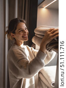 Smiling woman organizing clean towels on a shelf in warm morning light, captured in a cozy home interior with soft natural tones and casual clothing. Стоковое AI-изображение, AI-художник Катерина Бондарец / Фотобанк Лори