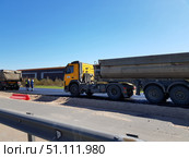 Road construction on a sunny day. In the foreground is a large yellow dump truck with a trailer used to transport construction materials. Next to it, workers in highly reflective work clothes are on the roadway. In the background, an industrial building and green bushes are visible. The road is partially closed and prepared for laying asphalt. In the foreground, there is a metal fence and temporary red plastic blocks used to fence off the repair area. The clear blue sky emphasizes the business and working atmosphere of the scene. (2019 год). Редакционное фото, фотограф Анна Кривицкая / Фотобанк Лори
