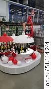 A festive display case in a supermarket with alcoholic beverages. In the foreground is a bright New Year's installation with bottles of Martini Asti and Martini Prosecco, designed as decorative Christmas tree balls in white and red. The composition is complemented by large plastic balls on the floor and lighting that creates a glowing effect. In the background, shelves with other drinks are visible, including beer and juices. The atmosphere of pre-New Year shopping and a festive mood in the store. (2018 год). Редакционное фото, фотограф Анна Кривицкая / Фотобанк Лори
