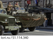 Military equipment from the Second World War on parade. In the foreground, you can see a DUKW amphibious transport in protective green paint with ropes attached to the hull. Next to it is an army truck from the same era. On board the amphibious vehicle are people in Soviet uniforms, some waving to the spectators. The vehicles move along a city street past a hotel building with Russian flags hanging above it. The atmosphere is that of a parade, a reenactment, or a memorial event dedicated to military history and the feat of soldiers. (2015 год). Редакционное фото, фотограф Анна Кривицкая / Фотобанк Лори