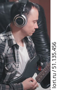 A musician in a cozy studio setting is focused on playing an electric guitar. He wears headphones and sports a plaid shirt over a white tee. Стоковое видео, видеограф Воловодов Владимир / Фотобанк Лори