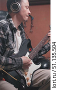 A musician plays a black electric guitar, wearing headphones. The cozy studio setting features a warm backdrop, emphasizing a creative atmosphere. The process of recording electric guitar in studio. Стоковое видео, видеограф Воловодов Владимир / Фотобанк Лори