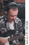 A musician plays an electric guitar while wearing headphones in a cozy, dimly lit room. The plaid shirt adds a casual vibe to the creative atmosphere. Стоковое видео, видеограф Воловодов Владимир / Фотобанк Лори