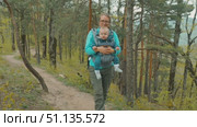 A woman smiles while carrying her baby in a backpack carrier. They walk along a dirt path in a serene forest setting filled with green pine trees and colorful undergrowth. Стоковое видео, видеограф Воловодов Владимир / Фотобанк Лори