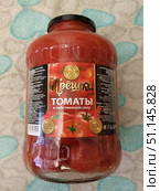 A large glass jar with pickled tomatoes in their own juice. The product is produced under the brand name "Pryoshka". The red label depicts juicy ripe tomatoes and the words "Tomatoes in their own juice", "GMO-free", "GOST". There is also a volume logo "3 liters" and information about the manufacturer on the sides. The jar is closed with a black metal lid. The background is light fabric with blue patterns. The photo conveys the image of a natural home-made product suitable for storage, cooking, winter preparations and traditional cuisine. (2019 год). Редакционное фото, фотограф Анна Кривицкая / Фотобанк Лори