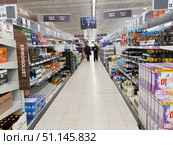 A long aisle in a large supermarket. The shelves on both sides of the aisle are filled with a variety of goods. On the left, you can see packages and cans of beer of various brands, including Heineken and other popular brands. On the right, there are drinks, cider, and large packages of household chemicals and washing powders. Above the shelves, there are signs in two languages: Finnish and Russian, indicating the categories "olut & siiderit" and "beer and cider". In the center of the aisle, you can see customers with carts. The interior of the store is done in light colors with a white floor and even lighting. The photo well conveys the atmosphere of everyday shopping and the wide range of goods in the supermarket. (2019 год). Редакционное фото, фотограф Анна Кривицкая / Фотобанк Лори