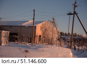 A winter rural landscape with a brick house, an electrical substation and wooden power lines. Snow covers the ground, trees and roof of the building, creating an atmosphere of peace and quiet. The frame shows footprints in the snow and dry plants protruding from under the snowdrifts. The low winter sun paints the building and trees in warm golden shades, contrasting with the cold blue of the snow and sky. The scene conveys the atmosphere of the Russian hinterland in winter, a combination of nature, snow and rural infrastructure. Стоковое фото, фотограф Анна Кривицкая / Фотобанк Лори