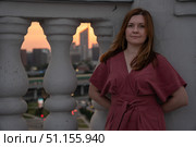 Woman in a rose dress leans against a rooftop balustrade at sunset with a blurred city skyline in the background. Стоковое фото, фотограф Софья Соломонова / Фотобанк Лори