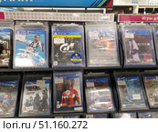A rack in an electronics and technology store displays video games for the PlayStation 4 console. The games are in transparent protective boxes with price tags. Popular titles can be seen in the foreground: FIFA 19, Gran Turismo Sport, Shadow Tactics, Tomb Raider, Hitman 2, FIFA 18 and Styx. The games are arranged in two rows, an upper and lower shelf. The covers depict recognizable characters and scenes typical of each genre - from sports simulators to action and strategy games. At the top of the shelf is the category "PS4 Games". The atmosphere of the store demonstrates the range of products for gamers. (2019 год). Редакционное фото, фотограф Анна Кривицкая / Фотобанк Лори