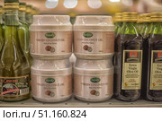 Shelves in a store with various types of vegetable oils. In the center of the frame are jars of 500 ml Baraka Virgin Coconut Oil in white plastic containers. To the left and right of them are bottles of Extra Virgin Olive Oil in green and dark glass containers. In the background, rows of similar products are visible, lined up on the display case. The photo is taken with an emphasis on the central row, creating the atmosphere of a grocery store. The image is suitable for illustrations on the topics of healthy eating, cooking, trade, product selection and the concept of organic food. (2015 год). Редакционное фото, фотограф Анна Кривицкая / Фотобанк Лори