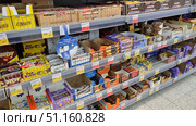 Supermarket display cases with a large selection of chocolate bars and sweets. Popular brands are neatly laid out on the shelves: Twix, Snickers, Nuts, Kinder Bueno, Picnic, as well as bars of lesser-known brands. The products are organized in boxes and sections with price tags, some of the packages are brightly colored - yellow, red, orange and purple. On the lower shelves are various candies and sweet snacks, on the upper ones - larger packages of chocolate. The photo illustrates the assortment of a grocery store, the theme of sweets, trade and consumer choice. (2021 год). Редакционное фото, фотограф Анна Кривицкая / Фотобанк Лори