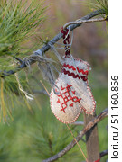 A mitten-shaped amulet, hand-made from coarse fabric (linen or burlap). The mitten is decorated with red thread embroidery in the form of a traditional Slavic ornament and decorative red braid with a wavy pattern. It is suspended on a tree branch with a red ribbon. The background is green pine needles and a softly blurred natural environment, which emphasizes the ethnic and ritual nature of the object. Such a talisman symbolizes protection, warmth and care, and is often used in folk traditions as a talisman. Стоковое фото, фотограф Анна Кривицкая / Фотобанк Лори