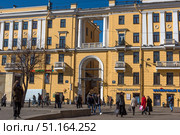 St. Petersburg's urban architecture on a sunny day. The image shows a bright yellow building in the Stalinist neoclassical style with white columns, balconies and arched openings. Below are shops, a jewelry store and a bank. Through the central arch, a passage with stairs and lighting is visible. In the foreground, people walk along a pedestrian street, some with bags and packages, creating the atmosphere of a busy city day. The architecture blends harmoniously with the blue sky and sunlight, emphasizing the brightness of the facade. (2019 год). Редакционное фото, фотограф Анна Кривицкая / Фотобанк Лори
