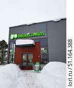Modern pet shop Musti ja Mirri on a winter day. The building is done in gray and red tones, the facade is decorated with a bright green sign with the image of a cat and a dog. In front of the entrance, high snowdrifts, neatly cleared paths and green street advertising of the store are noticeable. The brand logo is duplicated on the glass doors and windows. Snow falls slowly, creating the atmosphere of a northern winter. The photo reflects life in a cold climate and emphasizes the coziness of a modern pet shop for pets. (2019 год). Редакционное фото, фотограф Анна Кривицкая / Фотобанк Лори