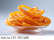 Traditional indian jalebi, a popular orange dessert made of deep fried batter soaked in sugar syrup, is presented on a glass bowl, ready to be enjoyed. Стоковое AI-изображение, AI-художник Юрий Поздников / Фотобанк Лори