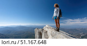 Female hiker with a backpack standing on the edge of a cliff, soaking in the breathtaking view of majestic mountain peaks under a clear blue sky on a sunny day. Стоковое AI-изображение, AI-художник Юрий Поздников / Фотобанк Лори