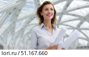 Young female architect smiling and holding blueprints while looking up in a modern building with glass ceiling, envisioning the future of construction. Стоковое AI-изображение, AI-художник Юрий Поздников / Фотобанк Лори
