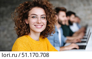 A cheerful young woman with curly hair and glasses smiles confidently while seated in a modern workspace. She is dressed in a vibrant yellow sweater, symbolizing positivity and warmth. Behind her, a row of focused individuals works on laptops, creating a sense of collaboration and contemporary professional culture. The image reflects modern workplace dynamics and empowerment, making it ideal for themes around Women's Day in Russia (March 8), education, technology, or youth-driven innovation. Стоковое AI-изображение, AI-художник Ксения Чунаева / Фотобанк Лори