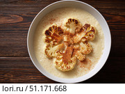 Cauliflower creamy soup with baked cauliflower florets on the top in a white bowl on a dark wood tabletop. Restaurant style dish. Hot, delicious plate of cream-of-cauliflower soup with melted cheese. Стоковое AI-изображение, AI-художник Марина Демидюк / Фотобанк Лори
