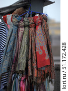 Bright oriental shawls and scarves hung on a rack for sale at a street market. The fabrics are decorated with complex ethnic-style ornaments: floral patterns, geometric elements, paisley and oriental motifs. The color scheme varies from rich blue and green to warm shades of red, orange and terracotta. The edges of the shawls are decorated with tassels. The atmosphere of the photo conveys the spirit of an oriental bazaar, handicrafts and ethnic fashion. Стоковое фото, фотограф Анна Кривицкая / Фотобанк Лори