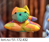 A soft toy in the form of a cat, made of bright fabric with a large checkered pattern. The toy is hand-sewn, has a simple shape and a minimalist design: ears, paws and a muzzle with black embroidered eyes. The colors of the fabric are orange, green, yellow, blue and purple, which gives the product a joyful and positive look. The toy is located on a blurred background, which makes it the main accent of the composition. Such an item can be used as a children's toy, a souvenir or an element of decorative interior. Стоковое фото, фотограф Анна Кривицкая / Фотобанк Лори