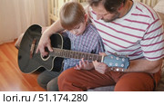 Handsome dad teaches his cute son to play the guitar sitting on sofa in the living room at home. Happy Father's Day. Father teaching his child to play guitar. Kid Learning music skill. Art education. Стоковое видео, видеограф Марина Демидюк / Фотобанк Лори