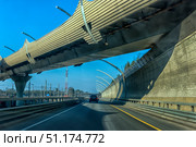 A modern section of a highway in St. Petersburg. The photo was taken from a car and shows an overpass with noise protection screens that rise above the road, creating a dynamic architectural perspective. In the foreground is a black car moving along perfectly smooth asphalt with yellow markings. On the sides are visible concrete fences, metal safety barriers and railway lines with power transmission line masts. In the background rises a red and white radio tower. The bright blue sky emphasizes modernity and open space. The atmosphere of the photo conveys the energy of the metropolis and the modern transport infrastructure of St. Petersburg. Стоковое фото, фотограф Анна Кривицкая / Фотобанк Лори