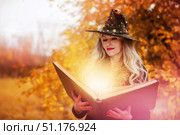 Halloween a woman with a magic spell book in a witch hat is conjuring, performing witchcraft over the autumn background of the forest. Стоковое фото, фотограф Лазаренко Светлана / Фотобанк Лори