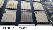 A stand in a store with samples of heating radiators. Various models of sectional batteries are fixed to the wall - bimetallic and aluminum radiators in white and gray. Each sample has a serial number (6, 7, 8, 10, 11, 12), which makes it easier for customers to choose. Radiators differ in the number of sections and design, some of them have additional stickers with information about the characteristics and the manufacturer. On the right, metal towel warmers are visible. The photo illustrates the range of goods for heating and engineering systems in a specialized store. (2018 год). Редакционное фото, фотограф Анна Кривицкая / Фотобанк Лори