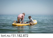 Two kids kneel on paddleboard, preparing for water fun and learning to paddle. Summer holiday activity with energy, joy, and playful mood. Summer vacation concept. Стоковое фото, фотограф Енваев Андрей / Фотобанк Лори