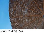 A close-up of a straw parasol, shot from below against a bright blue sky. The umbrella is constructed of twigs attached to wooden or metal crossbars, forming a geometric pattern. The sky is partially visible through the cracks in the weave, creating a play of light and shadow. The photo conveys the atmosphere of summer, relaxation on the beach or resort, a feeling of warmth and tranquility. Стоковое фото, фотограф Анна Кривицкая / Фотобанк Лори