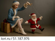 На фотографии представлена необычная и креативная постановка с элементами юмора и волшебства. Снегурочка в голубом зимнем костюме с белой меховой отделкой сидит на большом подарочном коробе, перевязанном красной лентой с бантом. Она держит в руках деревянный крест-марионетку и управляет маленькой фигуркой Деда Мороза, словно куклой на нитях. Дед Мороз в красном костюме с белым мехом и поясом улыбается и будто танцует, подчиняясь движениям. Атмосфера сюжета сочетает сказочность, иронию и праздничное настроение. Такой визуал станет необычным креативным баннером для рекламы, дизайна, рекламных кампаний, в соцсети, на сайт, для наружной рекламы, интернет-магазина и маркетплейса. Стоковое AI-изображение, AI-художник Sergey Starostin / Фотобанк Лори