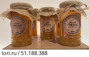 Rows of small, square glass honey jars with twine and decorative tags sit on a wooden surface, bathed in warm light; many more jars are visible,Assorted honey jars and candles are arranged on a table. Редакционное видео, видеограф Zita Khasanova / Фотобанк Лори