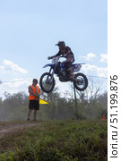 Magnitogorsk, Russian Federation - August 23, 2025: Male motocross rider in mid-air jump during enduro competition, with flagger signaling below, showcasing thrilling action and excitement in the sport. Редакционное фото, фотограф Ирина Ручушкина / Фотобанк Лори