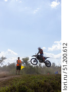 Magnitogorsk, Russian Federation - August 23, 2025: Young male motocross rider in mid-air jumps over a dirt track while a flagger watches, showcasing the excitement of enduro competitions. Редакционное фото, фотограф Ирина Ручушкина / Фотобанк Лори