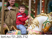 Young boy in red jacket enjoys a ride on a colorful carousel horse, accompanied by smiling woman during a fun outdoor outing. A child aged 3 years. Стоковое фото, фотограф Андрей Журавлев / Фотобанк Лори